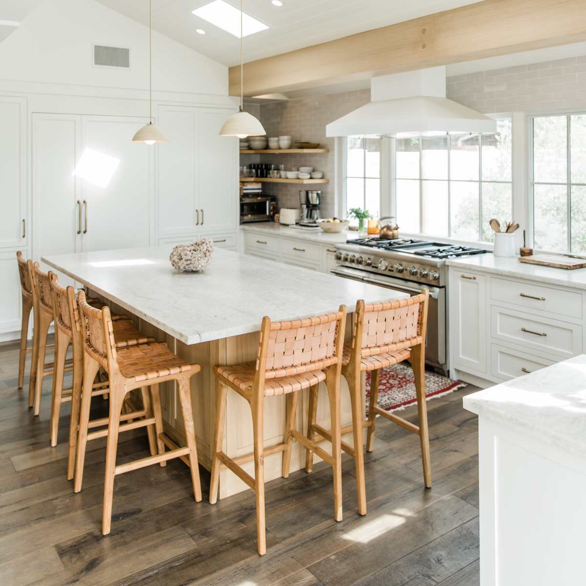A modern kitchen features a large marble island with woven chairs, stainless steel appliances, and plenty of natural light from skylights and windows.