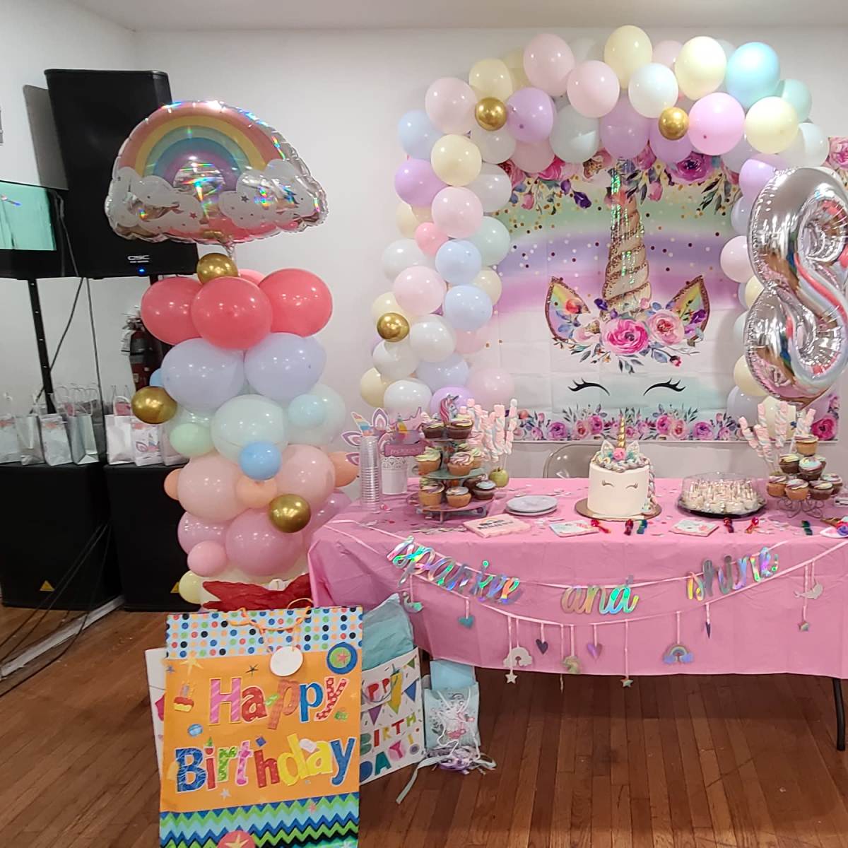 A birthday party setup features a table adorned with pink decorations, colorful balloons, and a large "8" balloon, set against a backdrop decorated with a cat illustration and a "Happy Birthday" banner.