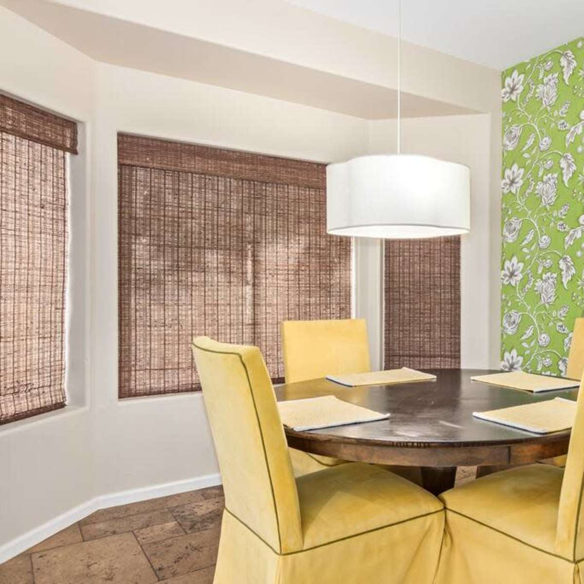 A round dining table with four yellow chairs is set in a room with a green floral-patterned accent wall and bamboo window shades.
