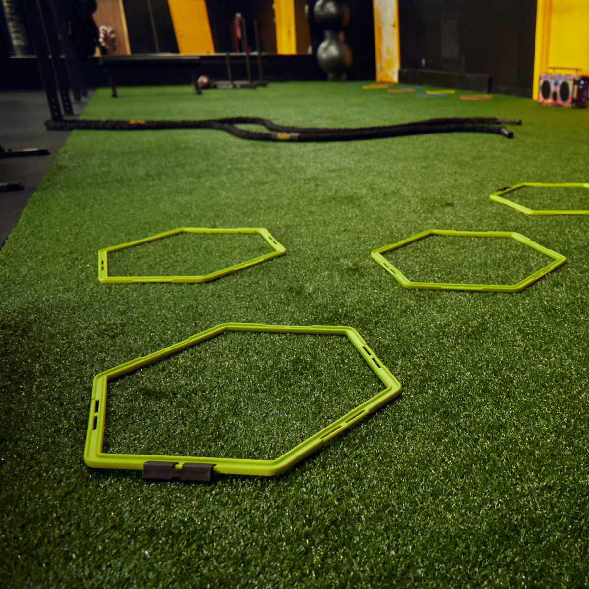 Five green hexagonal training cones are arranged on a synthetic turf surface in a gym setting.