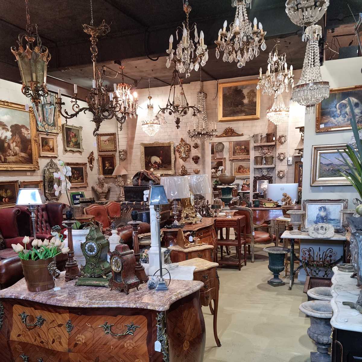 A vintage furniture store filled with ornate antiques, including chandeliers, paintings, and decorative items arranged on tables and shelves.