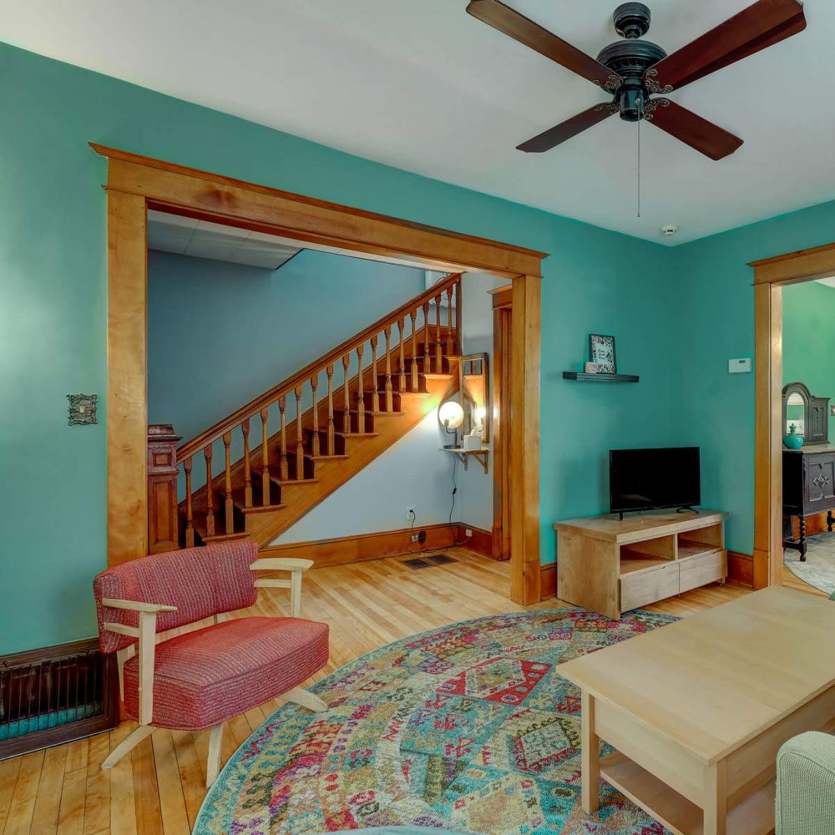 A living room features a teal wall, wooden trim, a ceiling fan, a coffee table, a couch, a chair, and a view of a staircase and dining area.