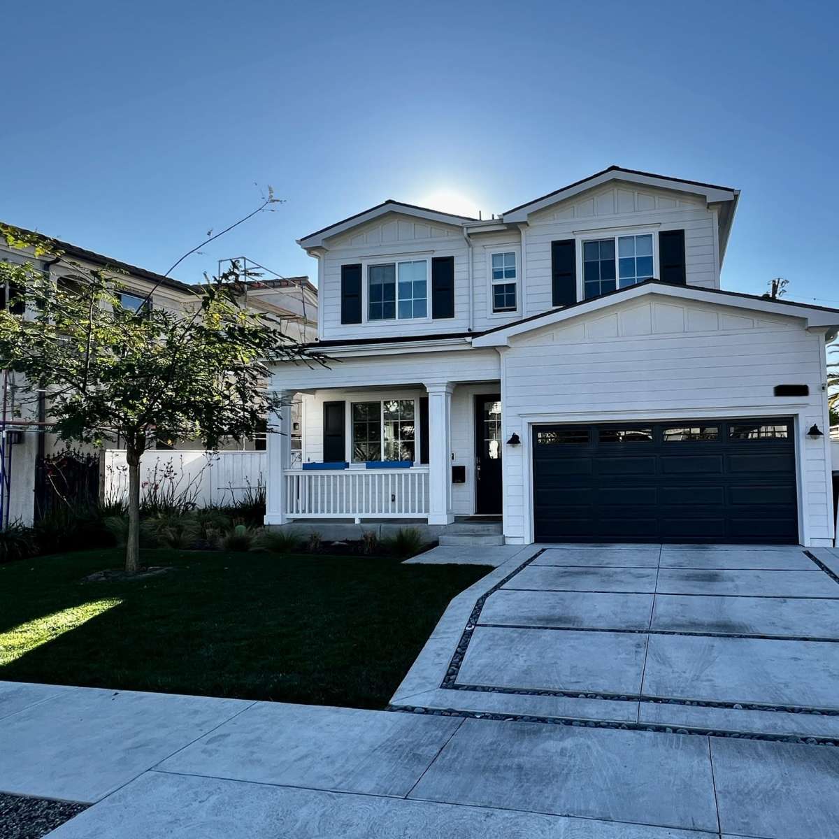 A two-story white house with a dark gray garage door and blue window shutters stands in a landscaped yard under a clear sky.