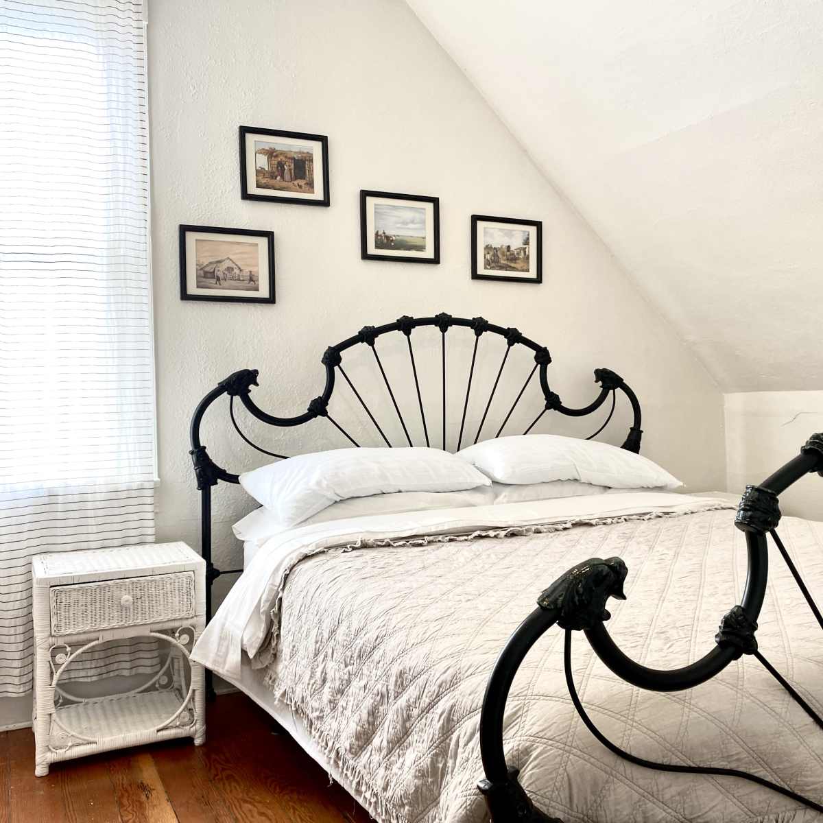 A bedroom features a black iron bed frame with white bedding, accompanied by a nightstand and framed pictures on the wall.