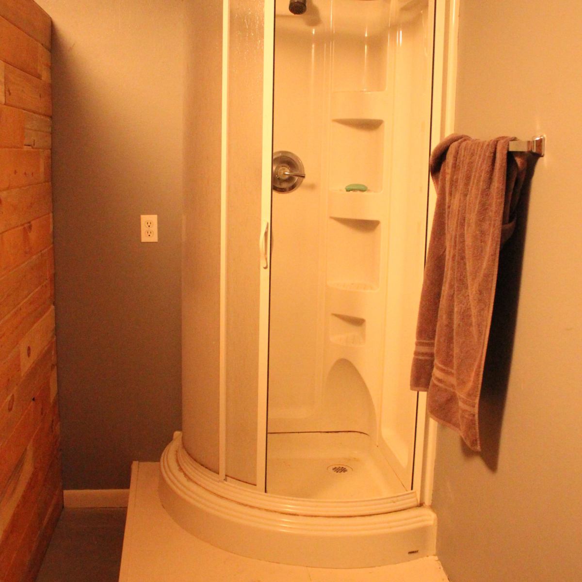 A corner shower with a curved glass door, situated in a bathroom with wooden paneling on one wall.