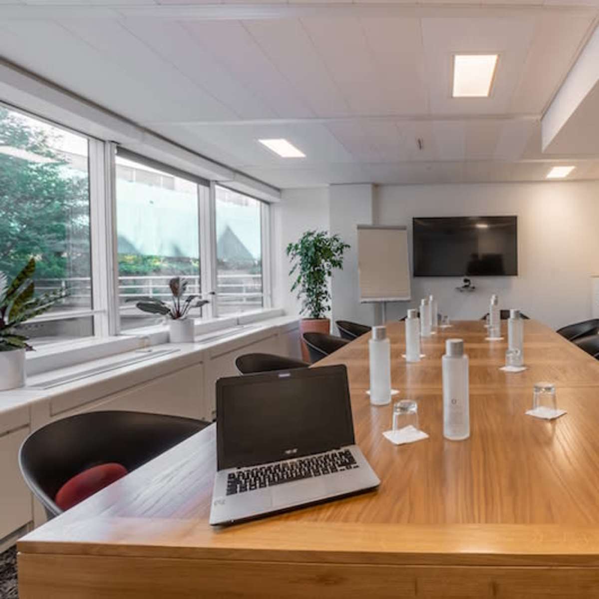 A modern conference room features a long wooden table surrounded by black chairs, with water bottles and glasses set for a meeting.
