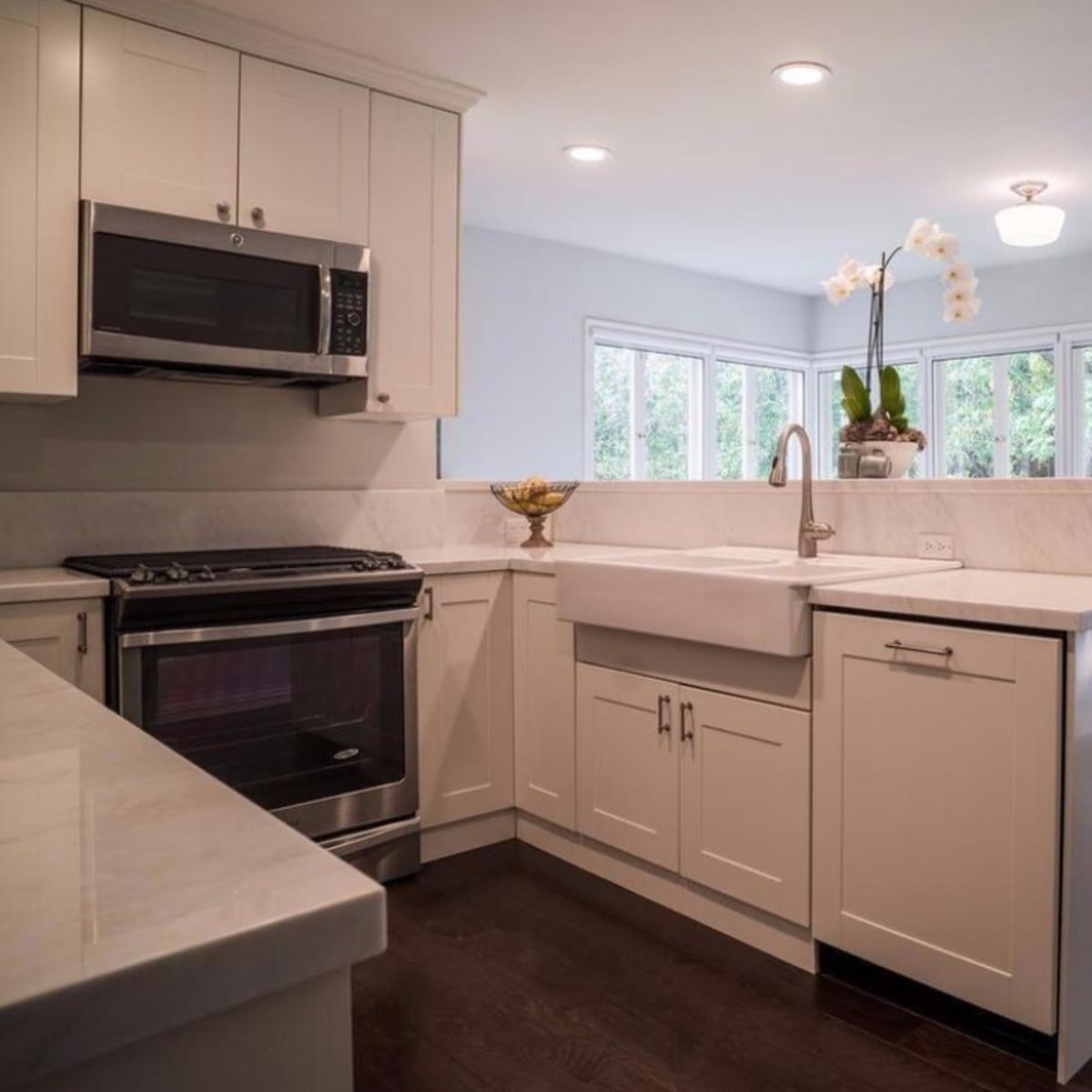 A modern kitchen features white cabinetry, a farmhouse sink, stainless steel appliances, and large windows overlooking a backyard.