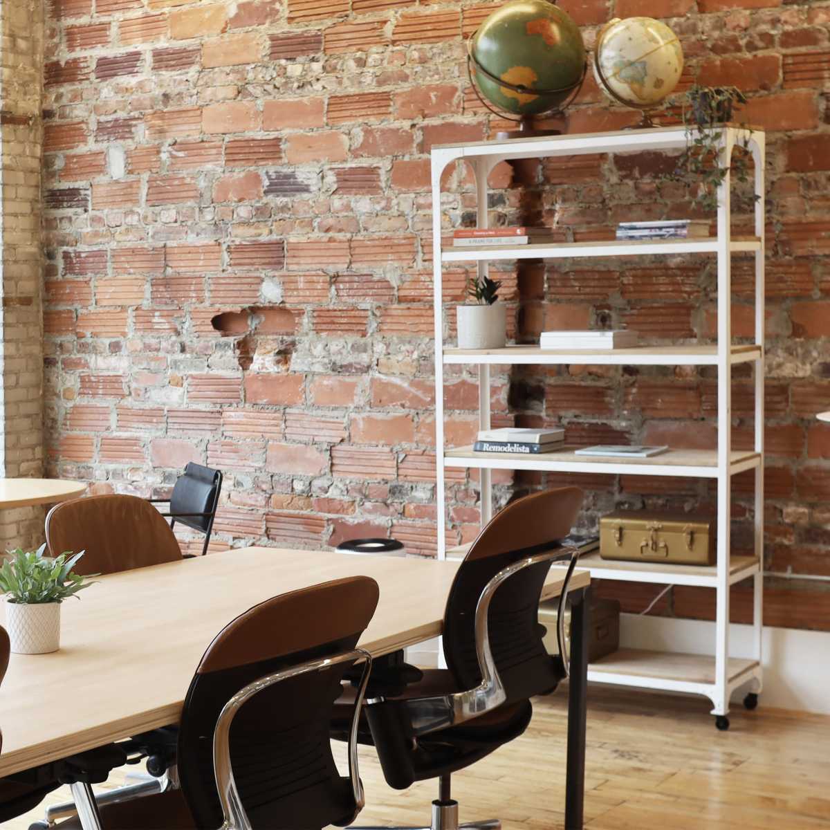 A modern meeting space with a wooden table surrounded by chairs, a brick wall in the background, and a shelf displaying plants and globes.