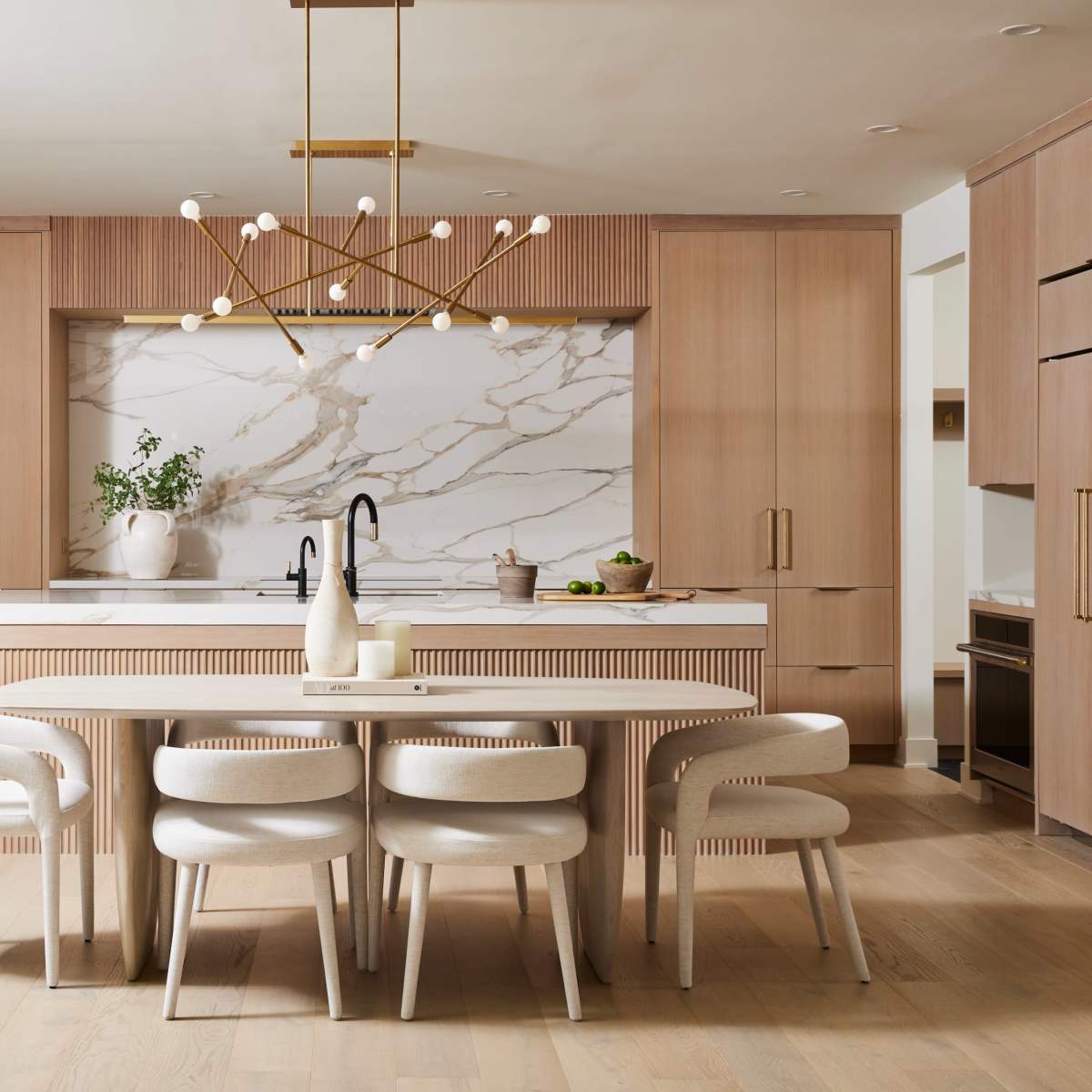 A modern kitchen features light wooden cabinetry, a marble backsplash, and a dining area with a round table and upholstered chairs.