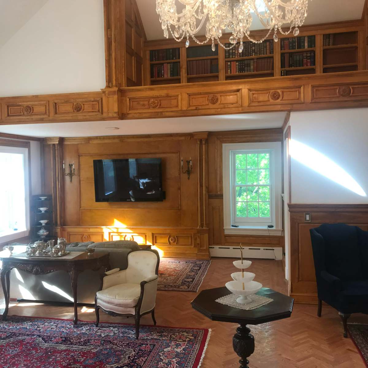 The room features a combination of wood paneling and large windows, with a chandelier above a seating area and a television mounted on the wall.
