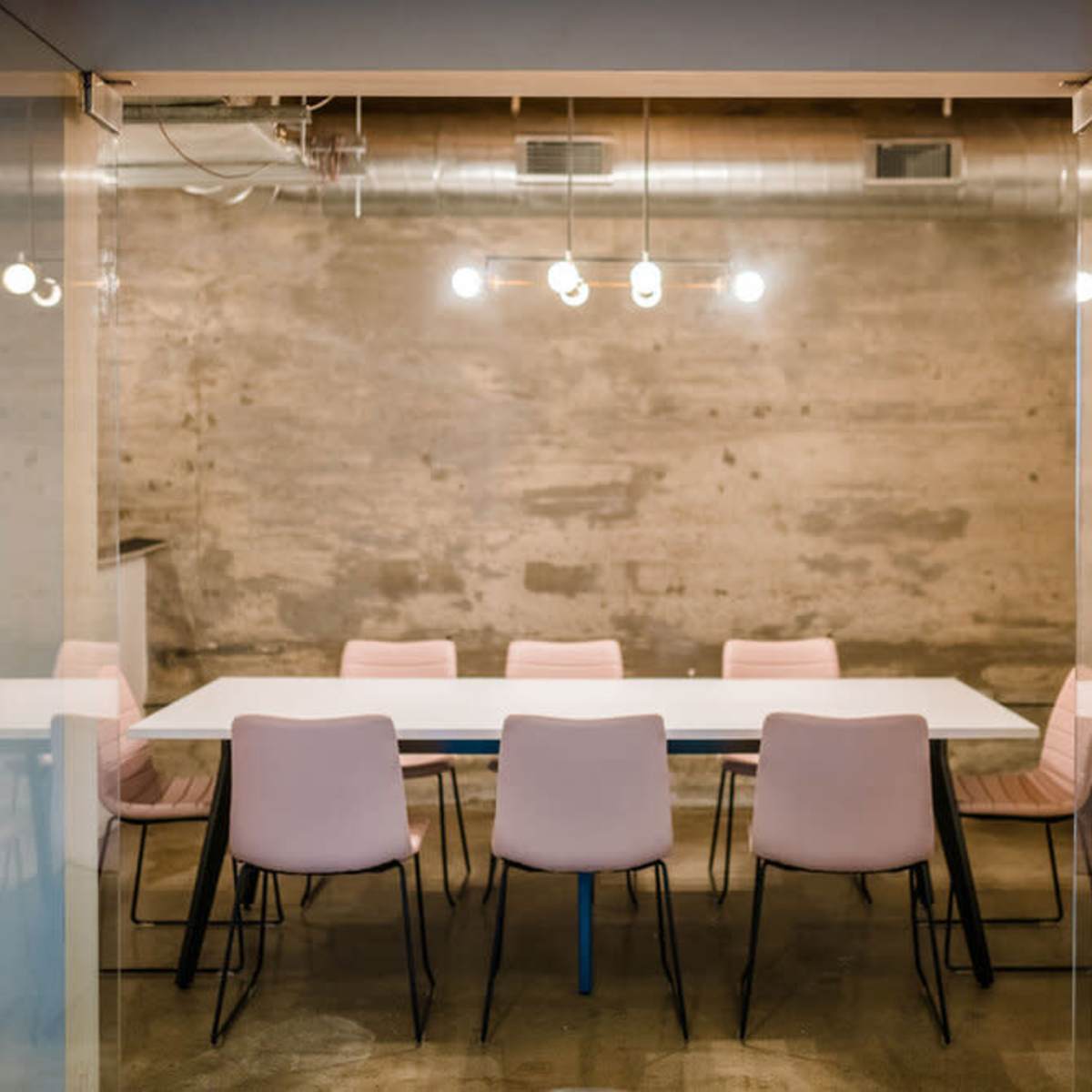 A minimalist conference room features a long white table surrounded by pink chairs under industrial-style lights.