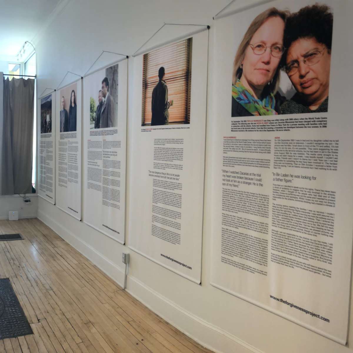 An indoor gallery space with several large framed photographs and text panels displayed on the walls, featuring a person with a dog near a table in the center.