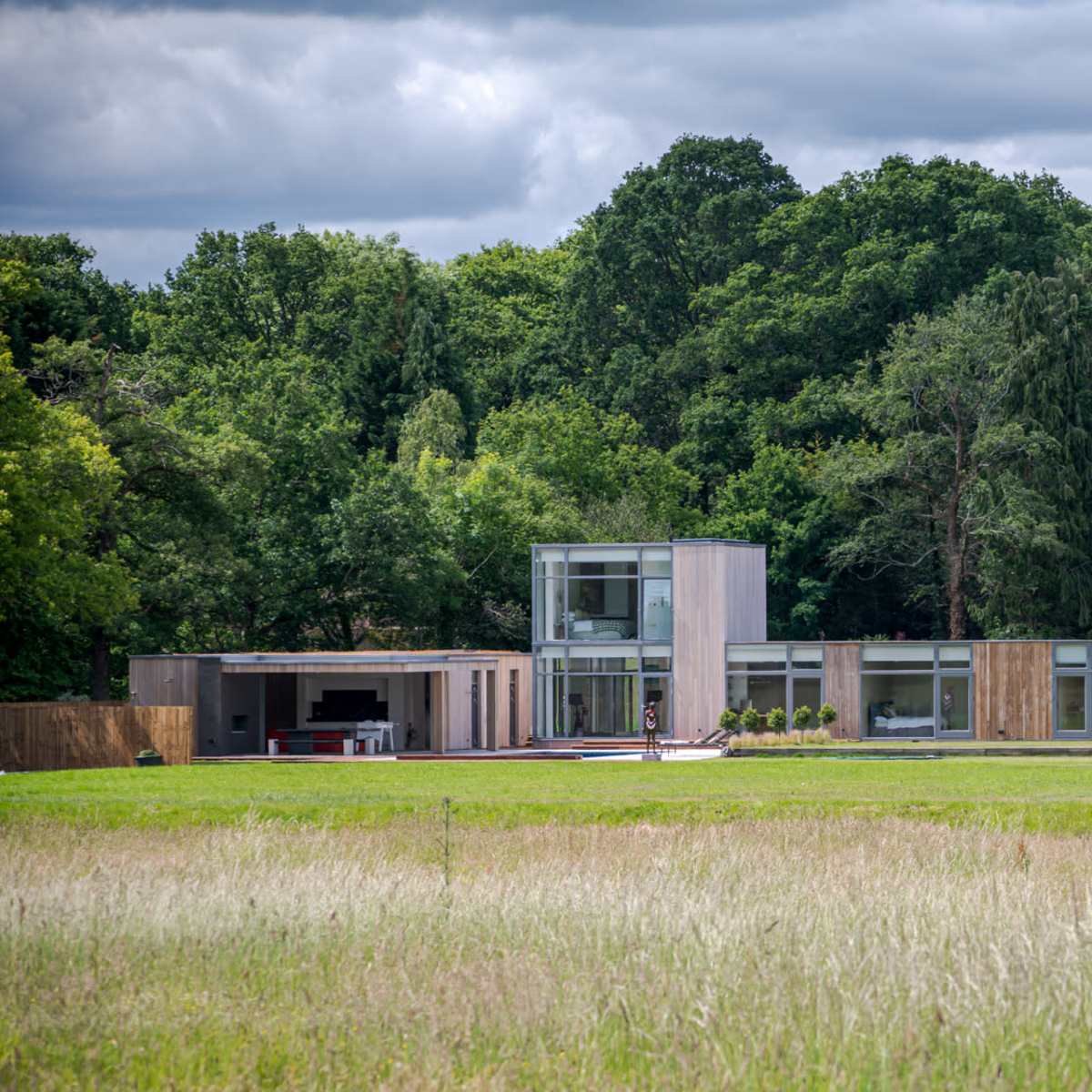 A modern, multi-level house with large glass windows is set against a backdrop of trees and a grassy field.