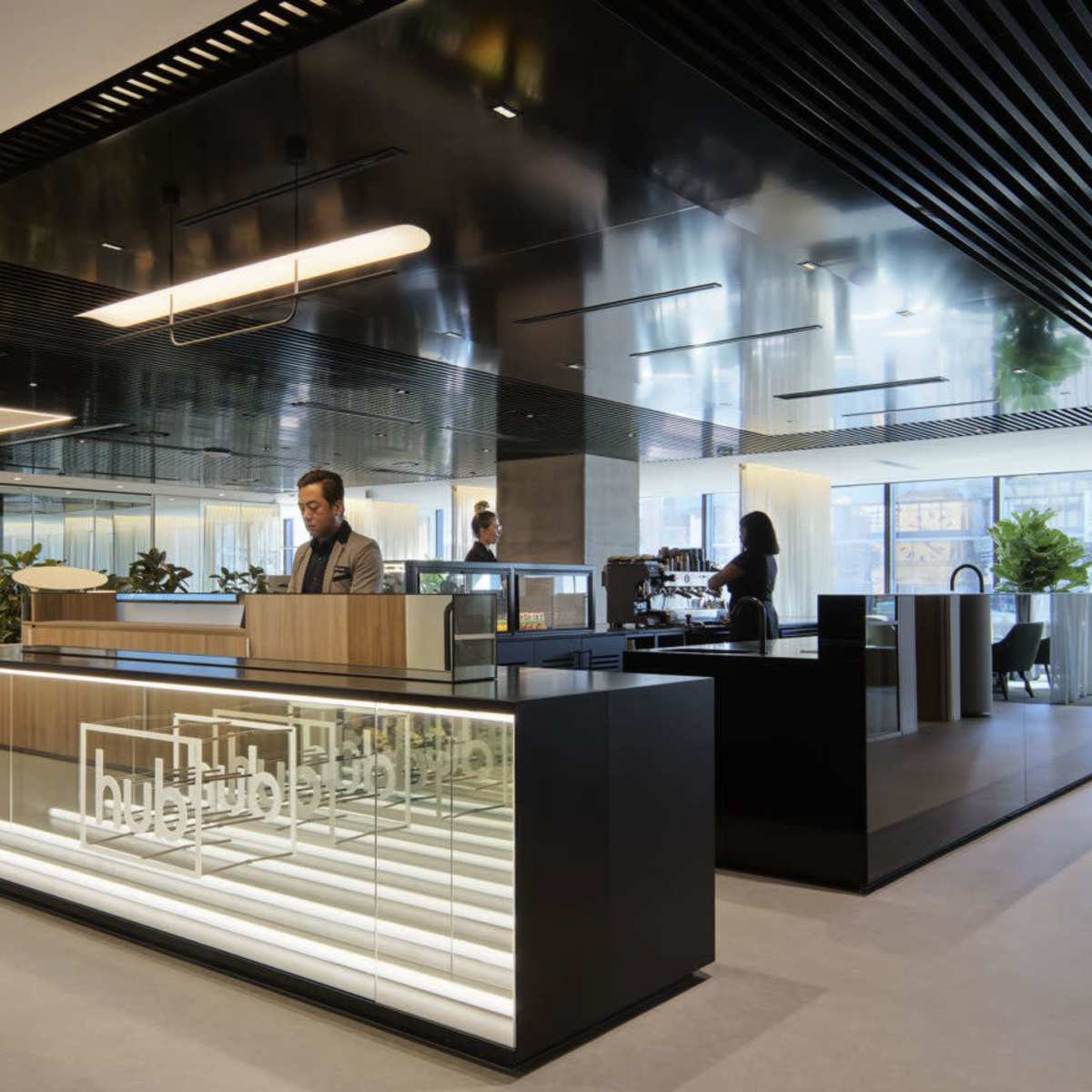 A modern office lobby with a reception area featuring a sleek desk and a staircase leading to an upper level, surrounded by greenery and large windows.