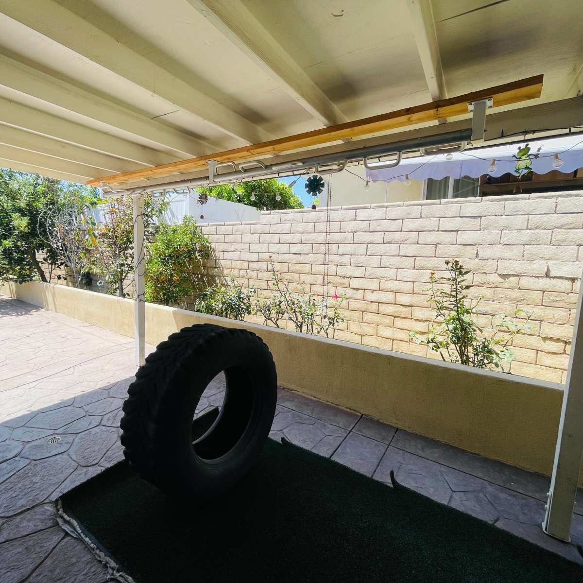 A view of a patio area with a large tire resting on a green mat and a garden against a stone wall in the background.