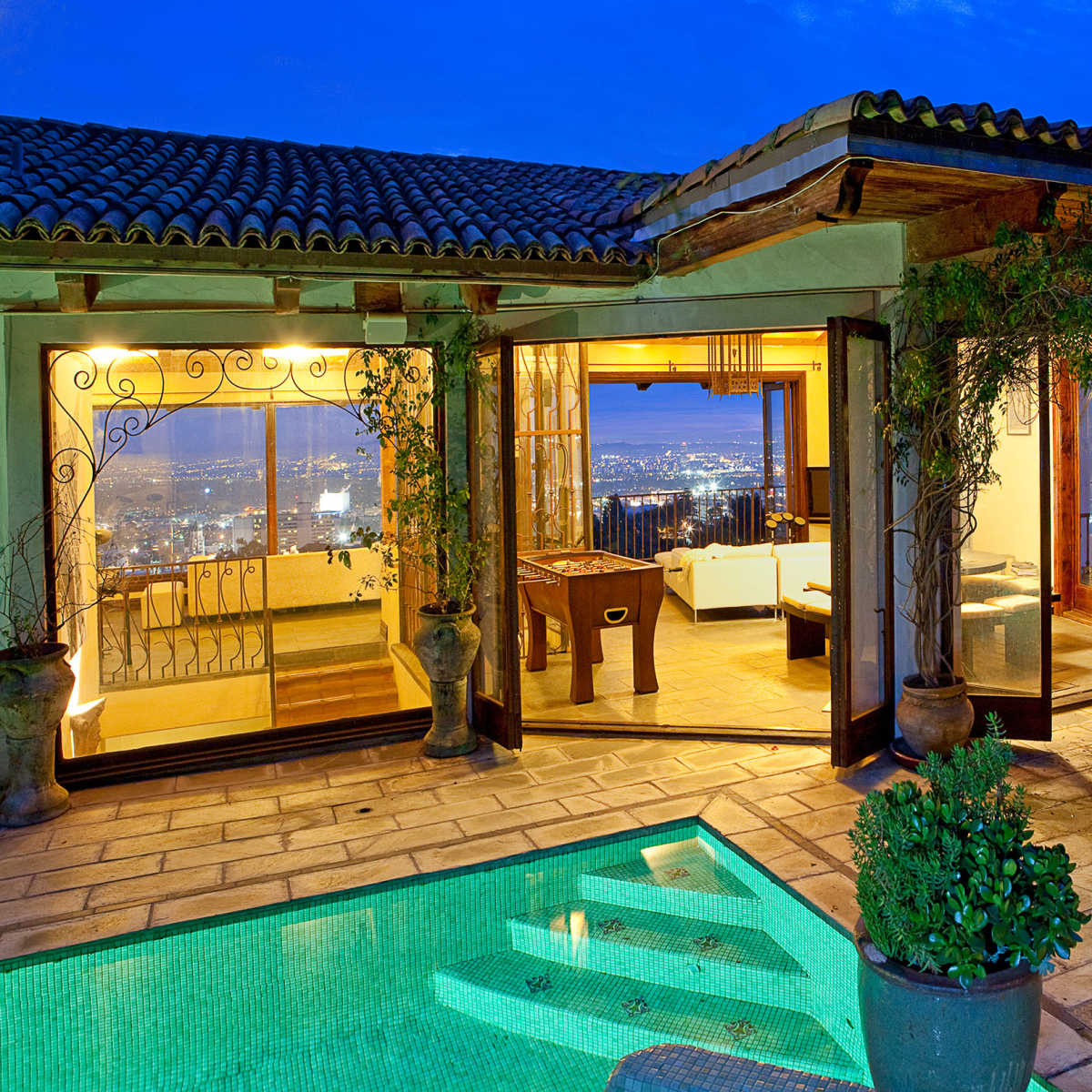 A luxurious villa with large glass doors opening to a pool area, featuring a view of a city skyline in the evening light.
