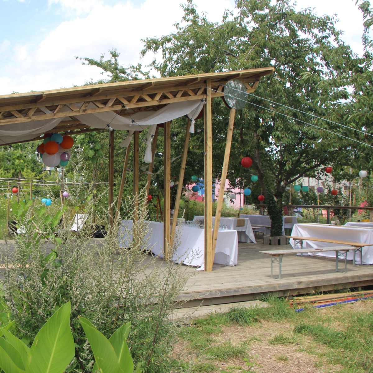 A wooden pavilion with a roof covered in fabric is set up in a garden, featuring tables and colorful decorations under trees.
