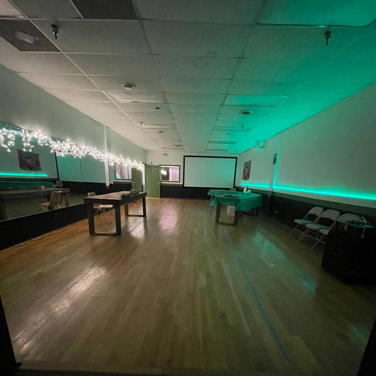 A spacious room with wooden flooring, decorated with string lights, and a projector screen at the end.