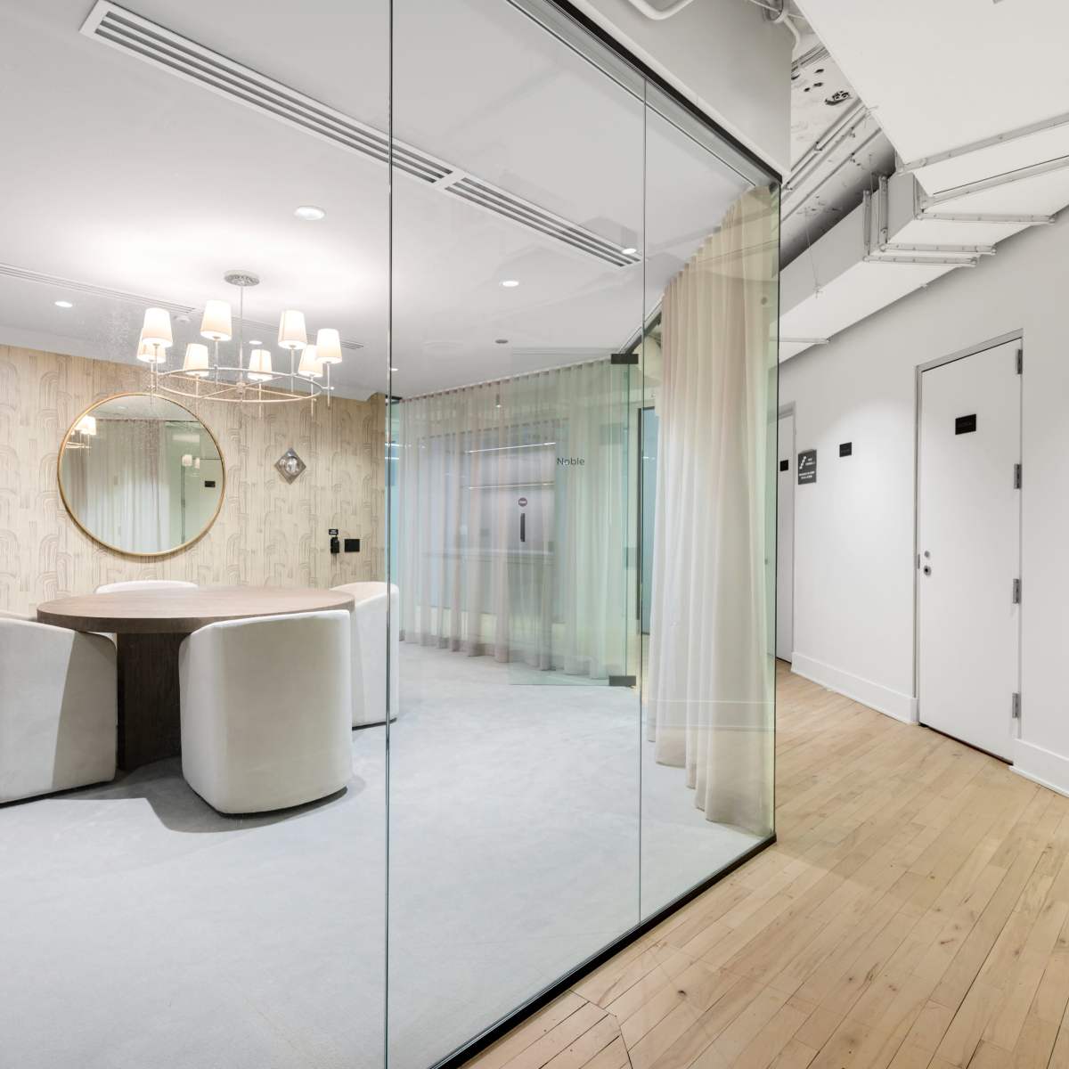 A modern meeting room features a round table surrounded by white chairs, with a large mirror and decorative lighting, enclosed by glass walls.