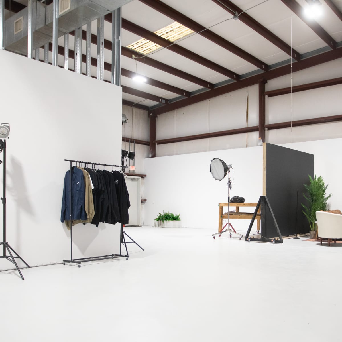 A spacious studio with white walls, a clothing rack, a seating area, and various lighting equipment arranged for a photoshoot.