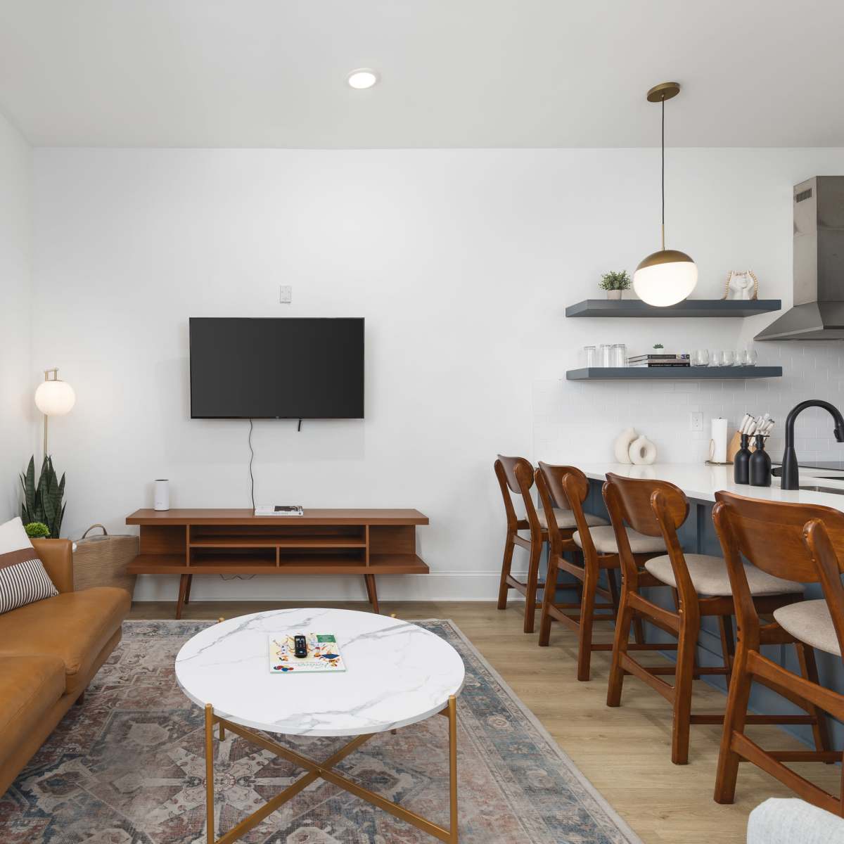 A modern living room features a brown leather sofa, a marble coffee table, and a wall-mounted television, with an adjoining kitchen area that includes wooden bar stools and shelves.