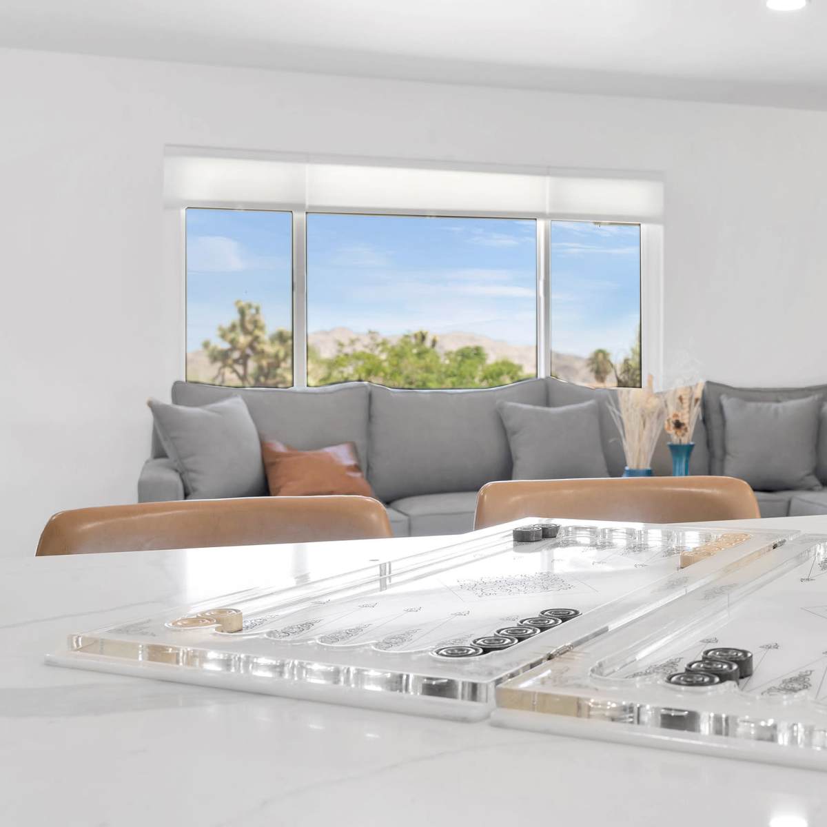 A game board is positioned on a marble table in front of a spacious seating area with gray cushions and a window showcasing an outdoor view.