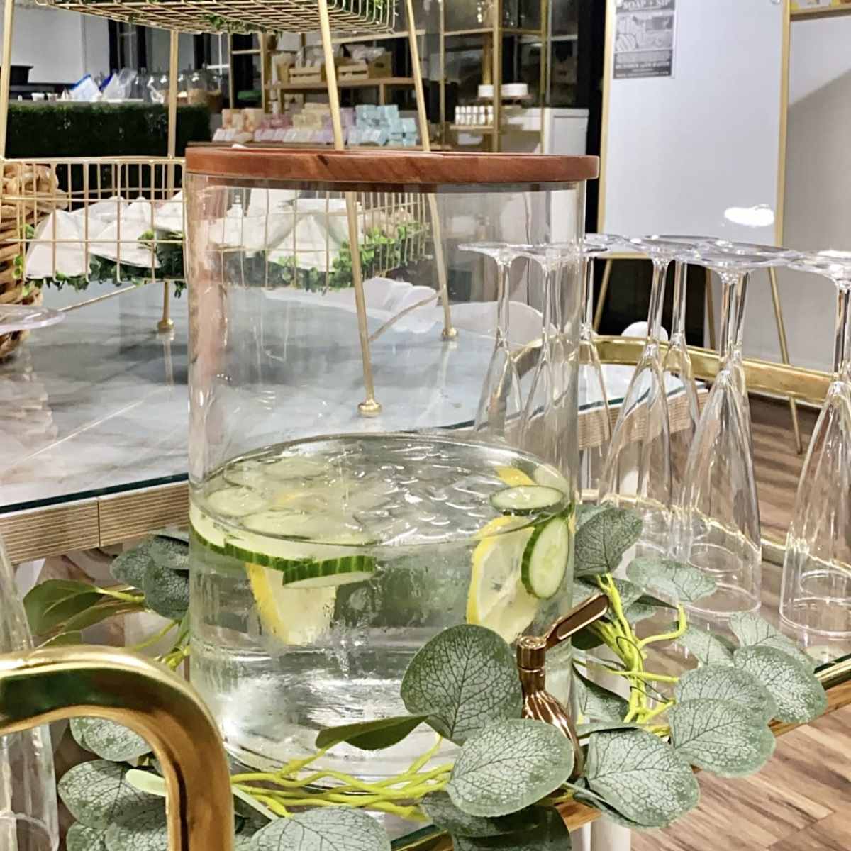 A glass container filled with water, lemon, and cucumber slices is displayed on a table surrounded by elegant glassware and greenery.