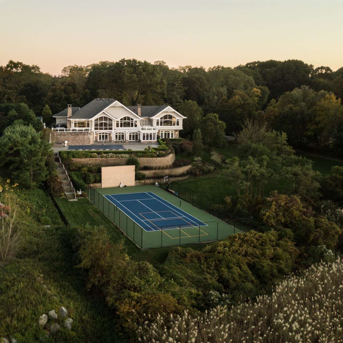 A luxurious house with multiple balconies overlooks a green tennis court surrounded by landscaping at sunset.