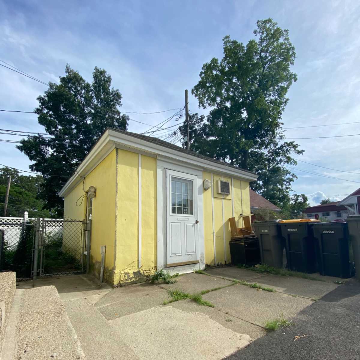 A small yellow shed with a white door and an air conditioning unit sits next to a pathway and several trash bins.