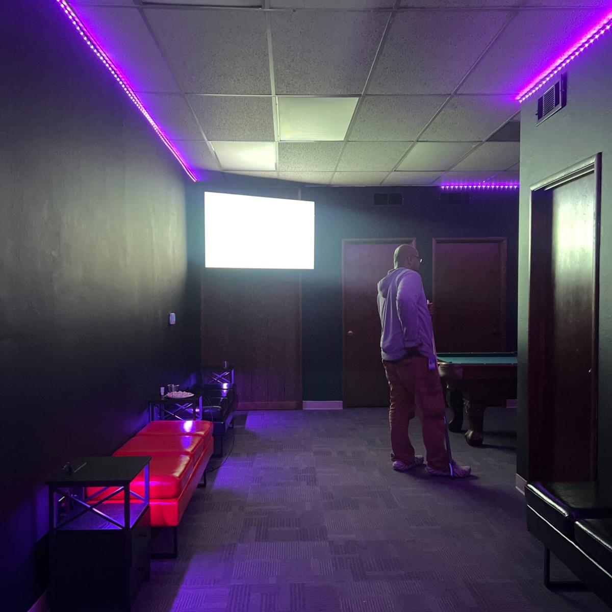 A man stands near a pool table in a dimly lit room with purple LED lighting and a blank screen on the wall.