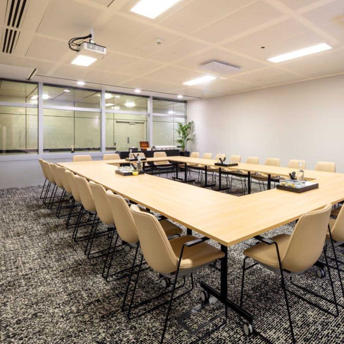 A large conference room features a rectangular table surrounded by beige chairs, with a projector and glass wall visible in the background.