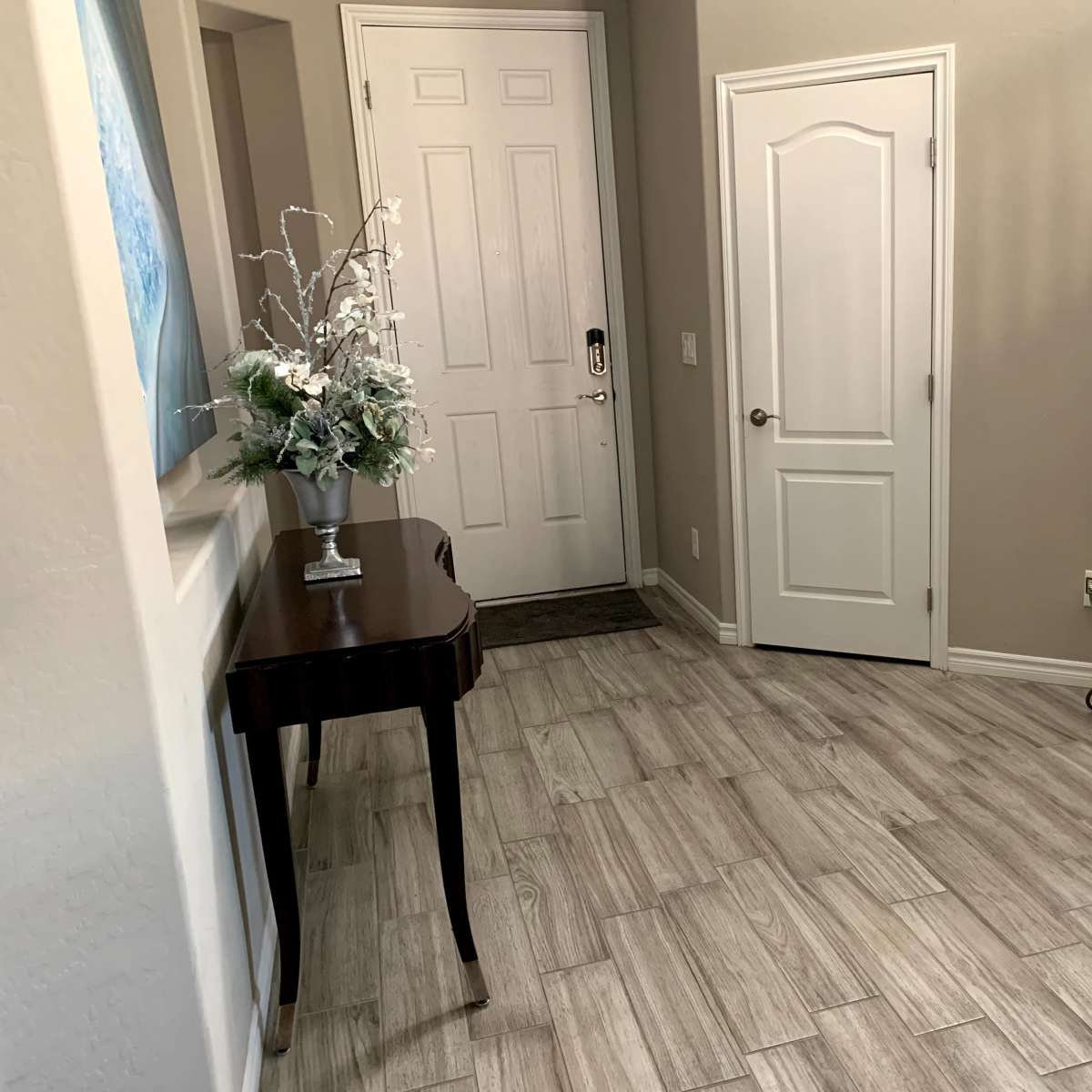 A hallway featuring a small table with a vase, a door leading outside, a closed interior door, a staircase, and a decorative mirror mounted on a stand.