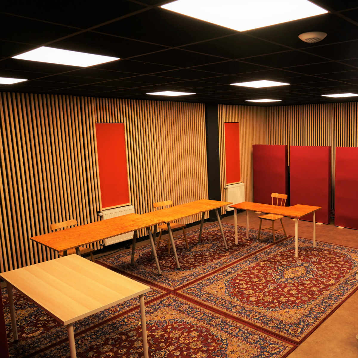 A room with several wooden tables arranged on a patterned rug, flanked by red window coverings and divided by red partitions.