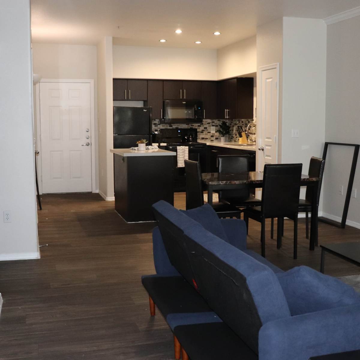 A modern living space with a blue couch facing a dining area and an open kitchen in the background.