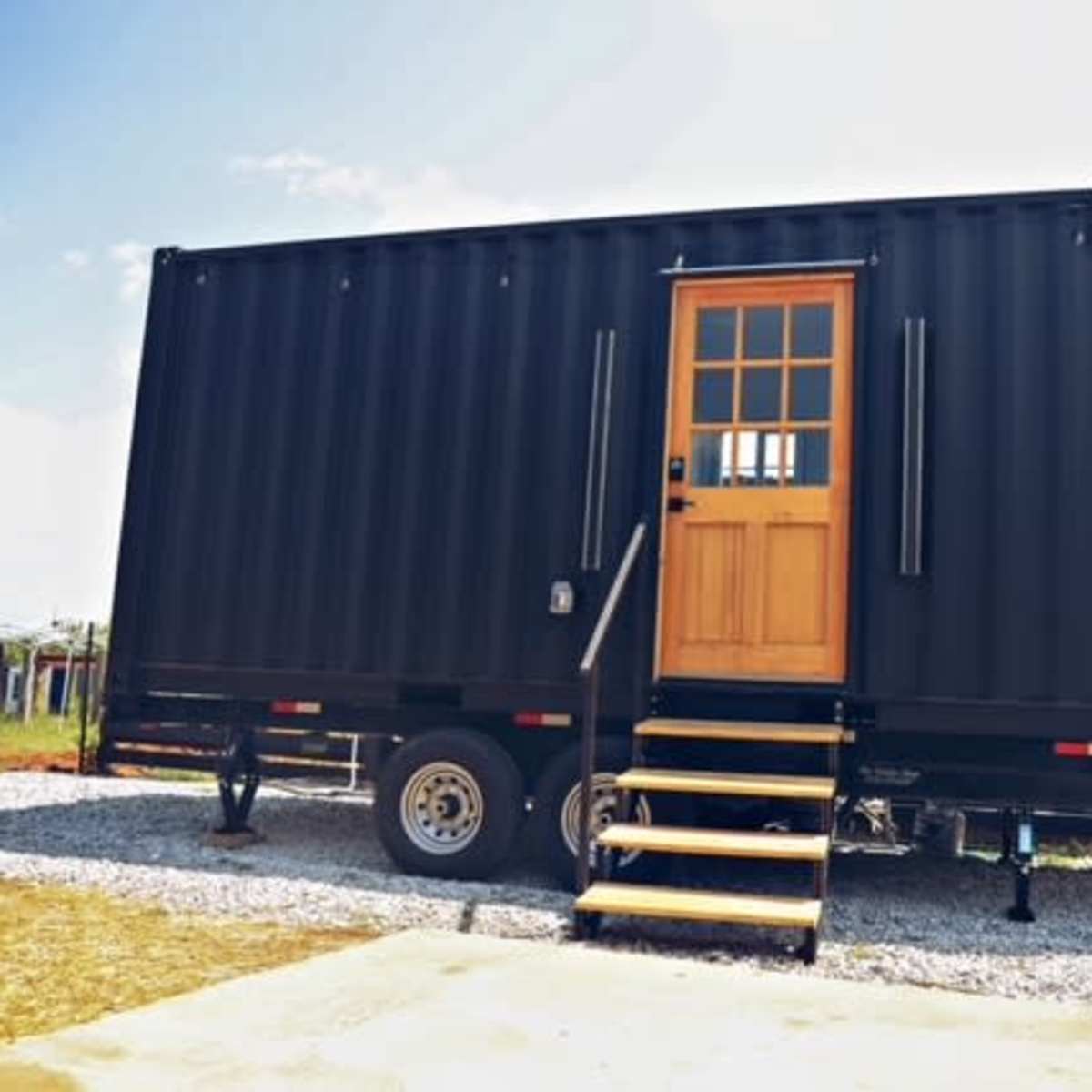 A black shipping container with a wooden door and steps sits on a gravel surface, surrounded by an open landscape.