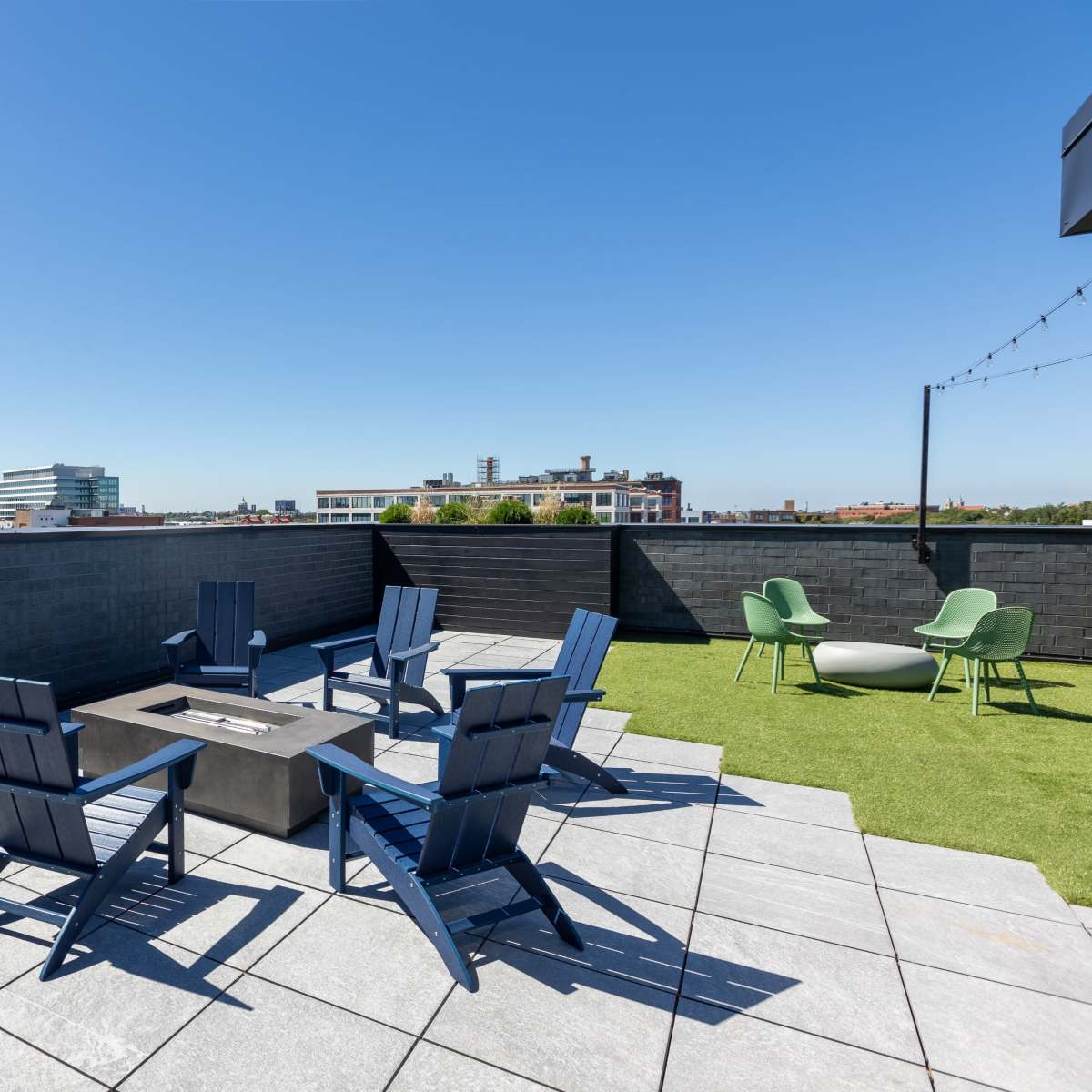 A rooftop seating area features several blue chairs surrounding a square fire pit on a paved surface, with green chairs nearby and a clear blue sky overhead.