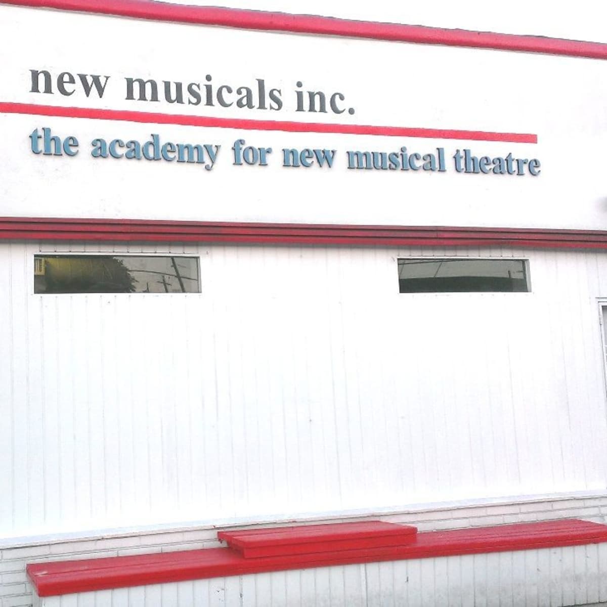 A white building with red trim featuring the sign for "New Musicals Inc." and "The Academy for New Musical Theatre" along with a red door labeled "5628."