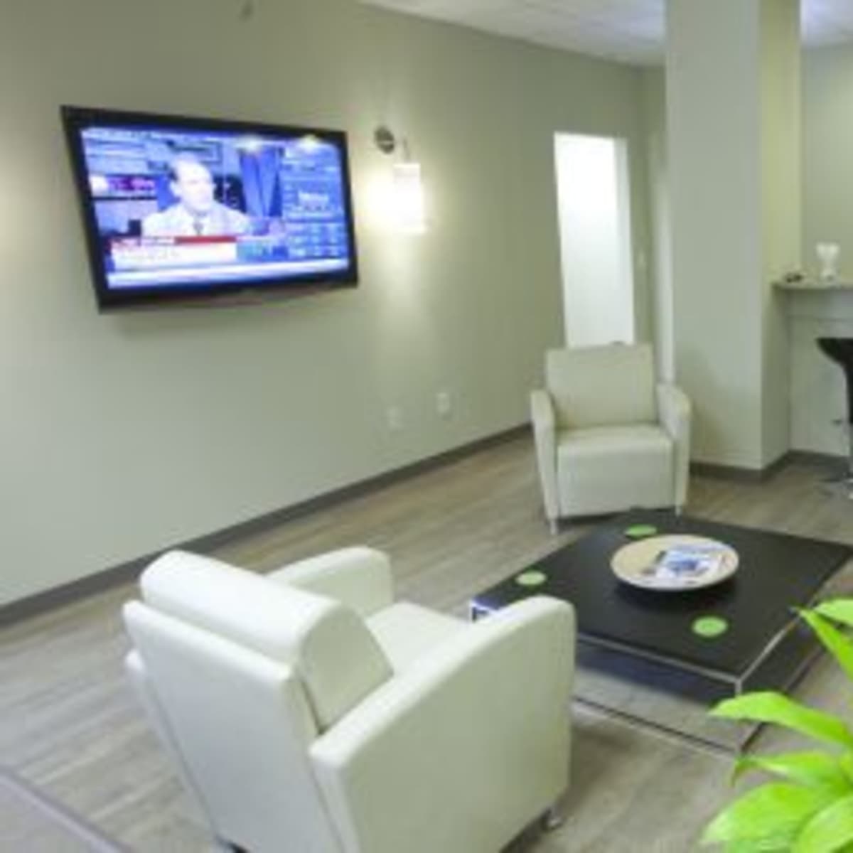 A modern waiting area with two white armchairs, a coffee table, a plant, a flat-screen TV displaying news, and a small bar with high stools.