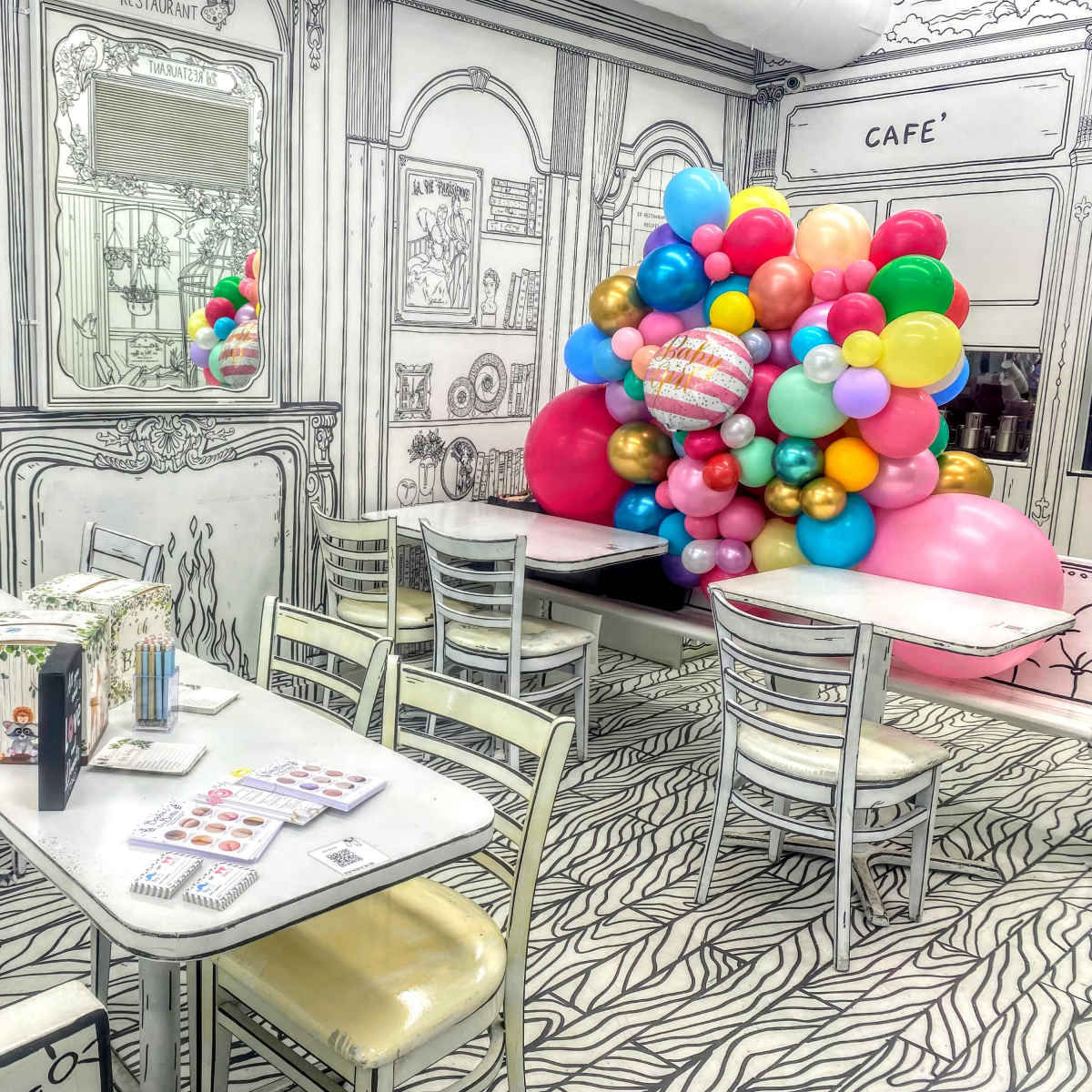 A whimsical café interior featuring outlined furniture and decorations, with colorful balloons arranged in a corner.