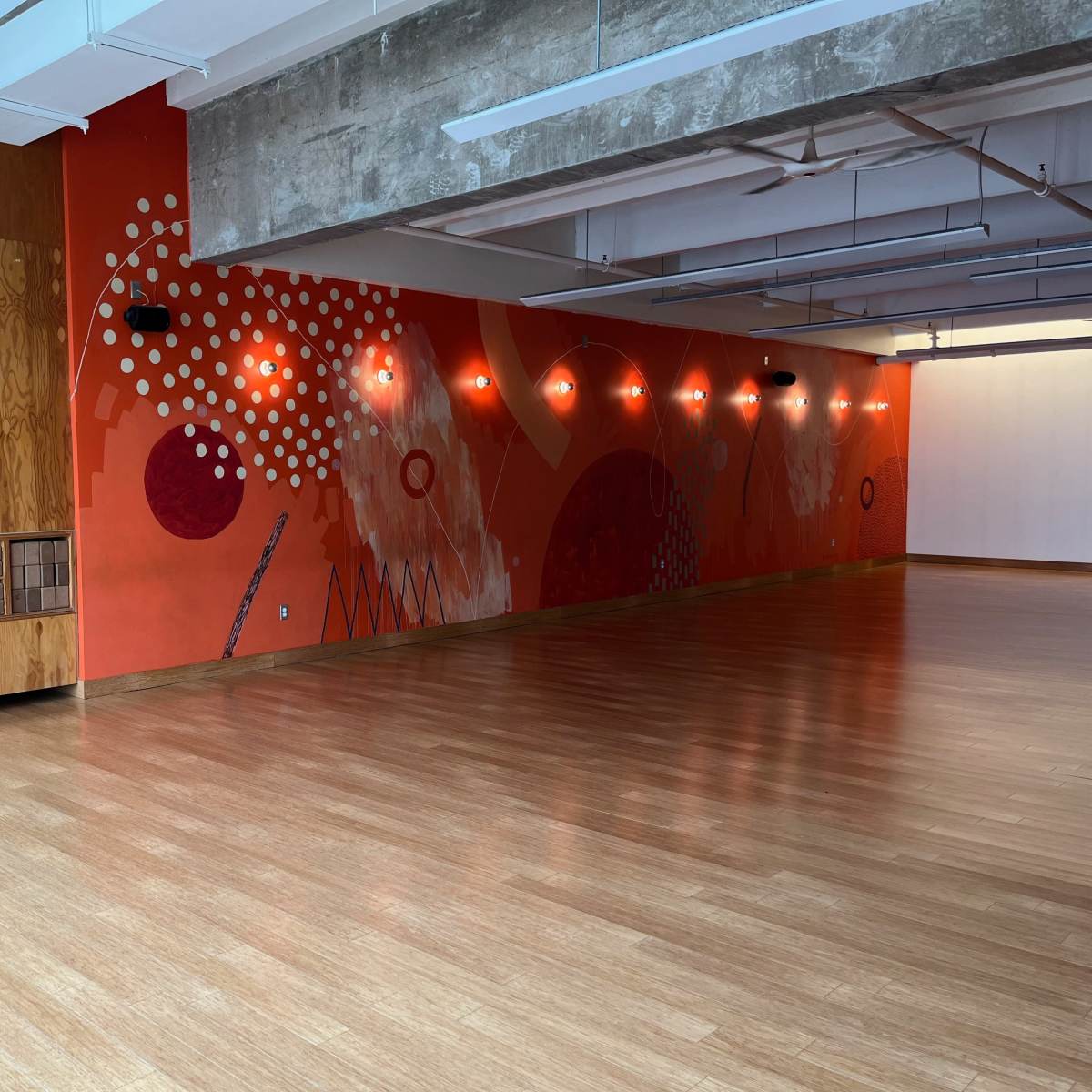 A spacious studio with wooden floors, a decorated orange wall featuring abstract designs, and shelves holding neatly stacked items.