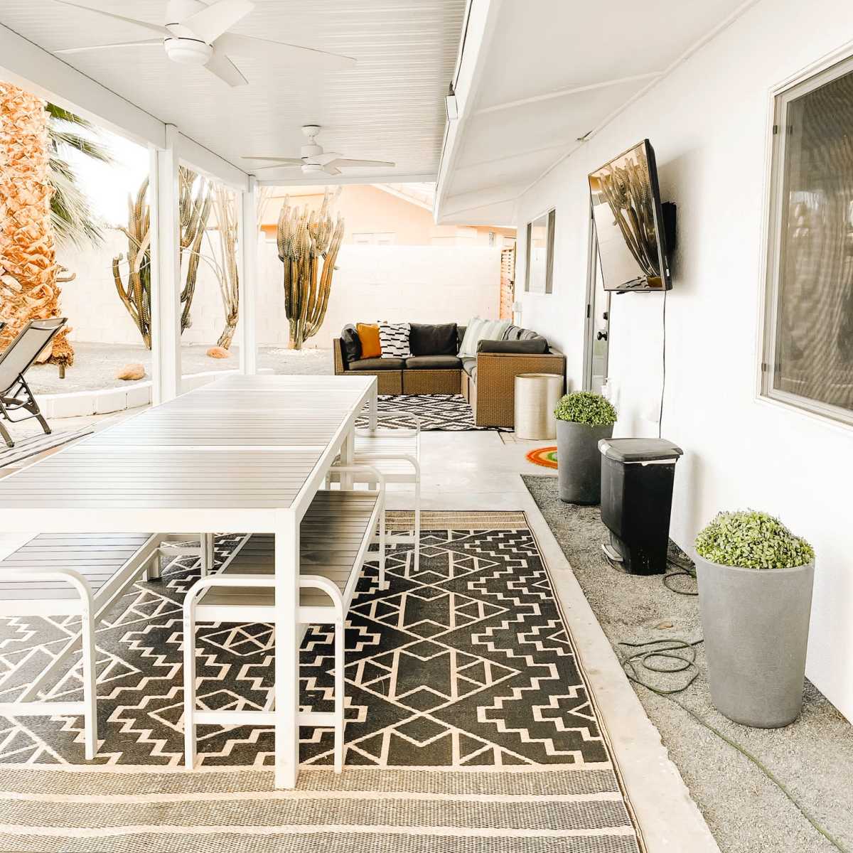 A covered outdoor patio with a dining table, comfortable seating area, and potted plants, all set against a backdrop of palm trees.
