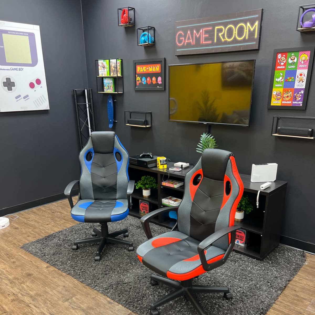 A game room featuring two office chairs, one blue and gray and the other red and gray, positioned on a dark rug in front of a TV and storage shelves displaying video game items.