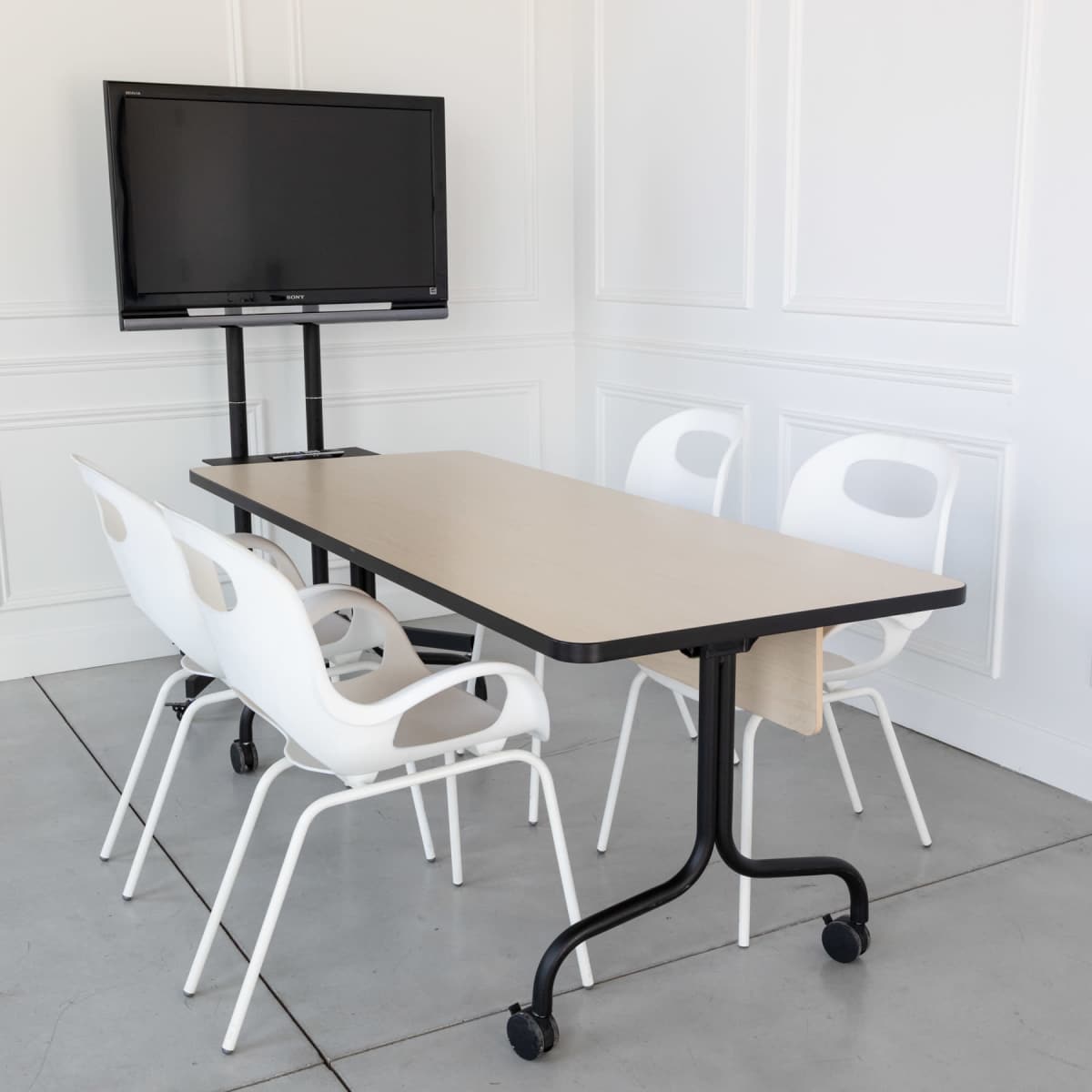 A modern conference room setup features a light wooden table with a black frame and four white chairs, alongside a mounted TV on a stand.