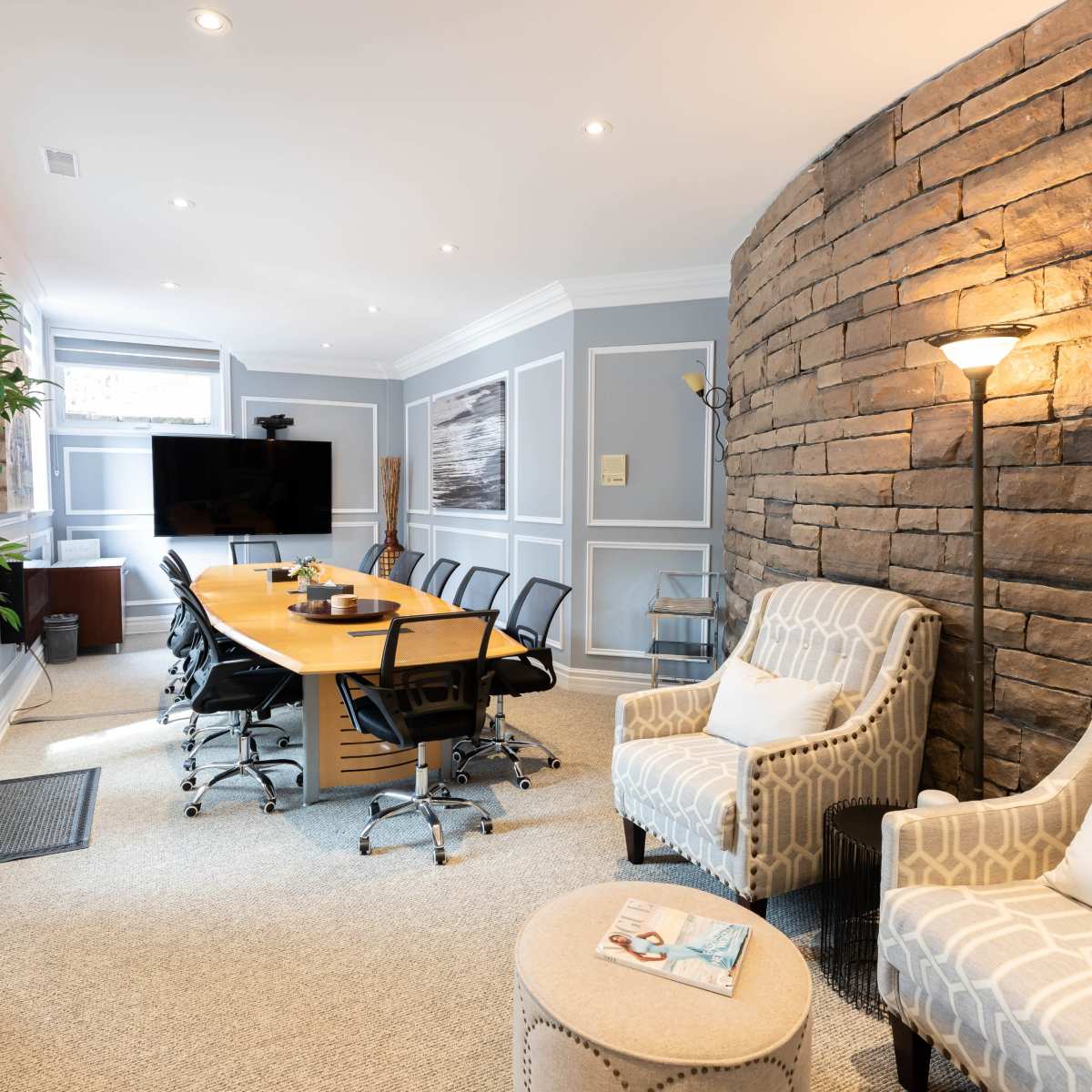A modern conference room featuring a long wooden table surrounded by black chairs, a TV screen on the wall, and comfortable seating with a stone accent wall.