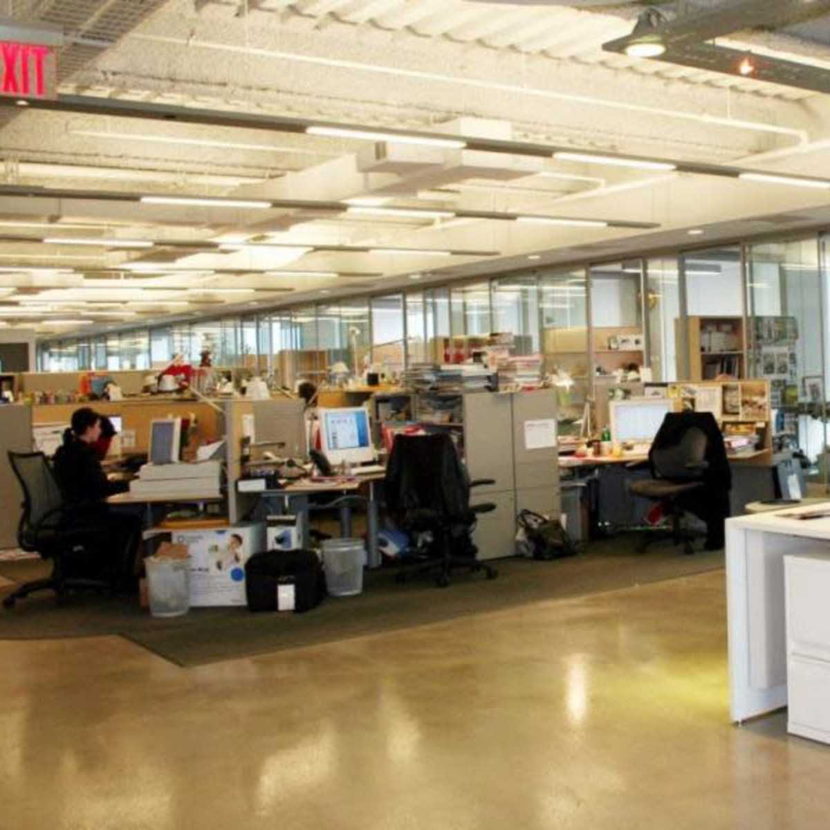 An open office space with cubicles, desks, computers, and large windows allowing natural light.