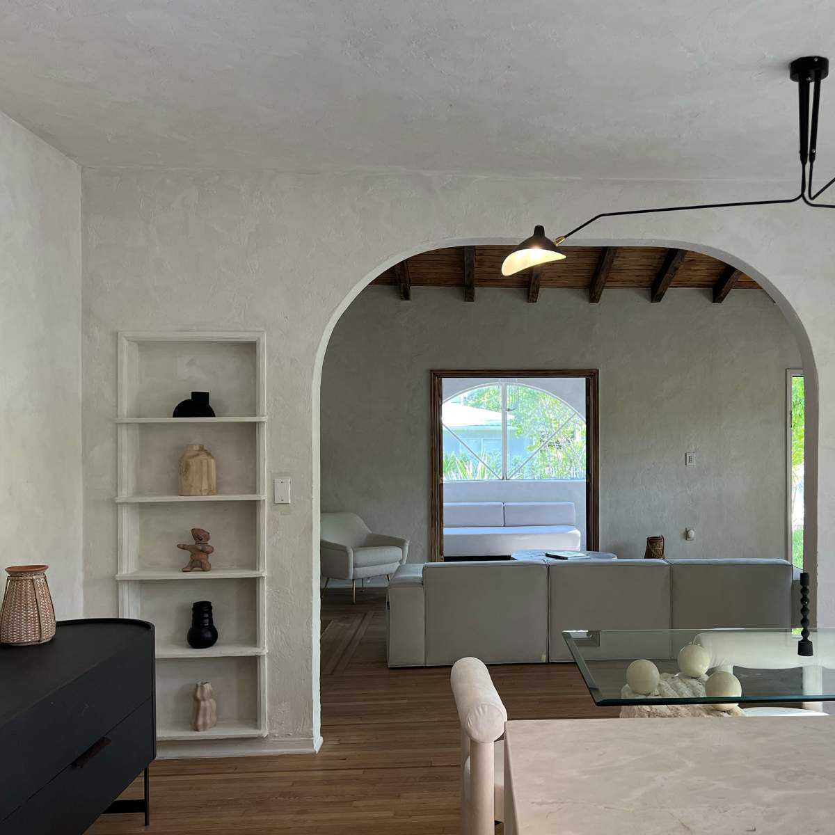A modern living space with a curved archway, minimalist shelves displaying decorative items, and a view of a furnished area beyond.