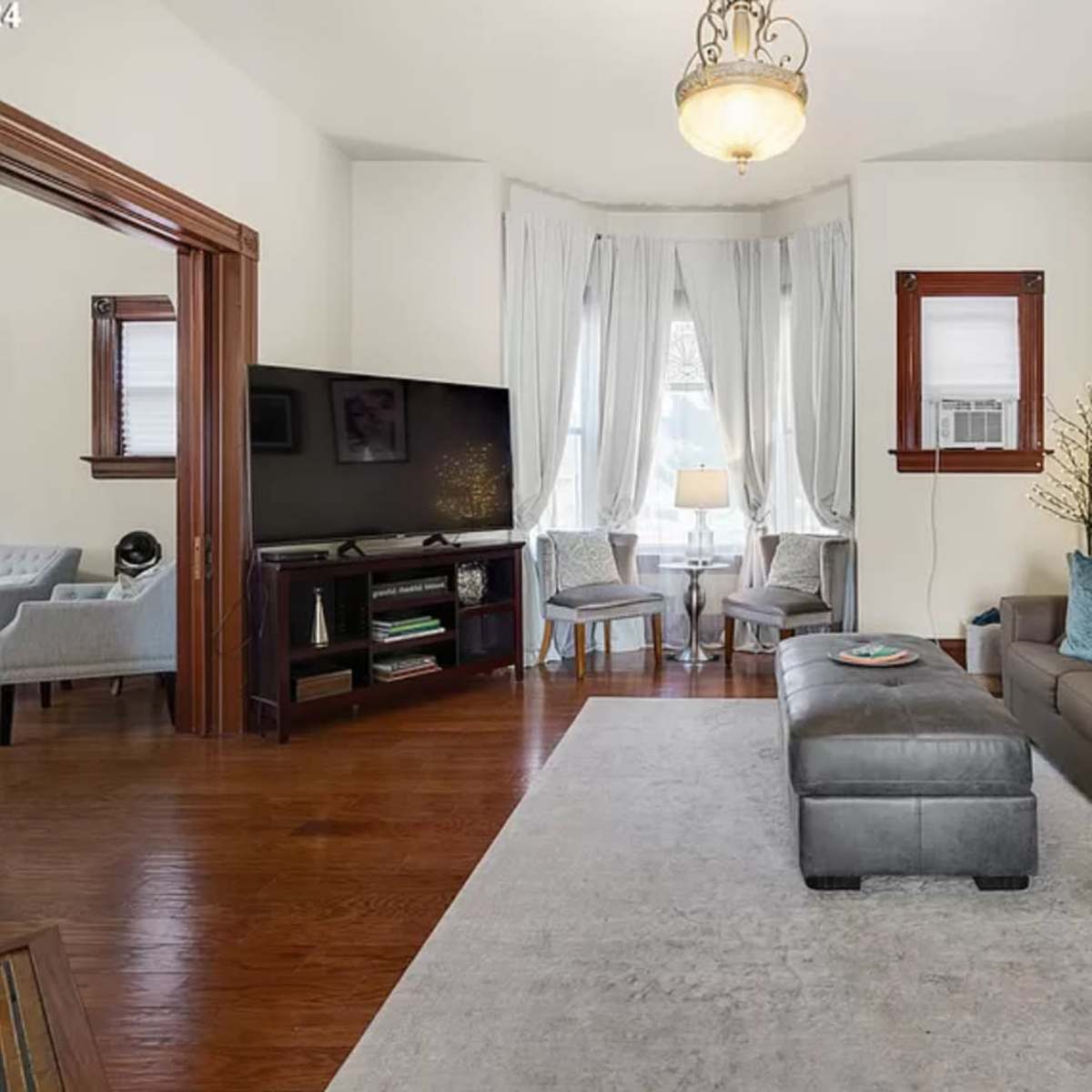 A living room features a gray sofa, an ottoman, a television, and two chairs near a window with sheer curtains.
