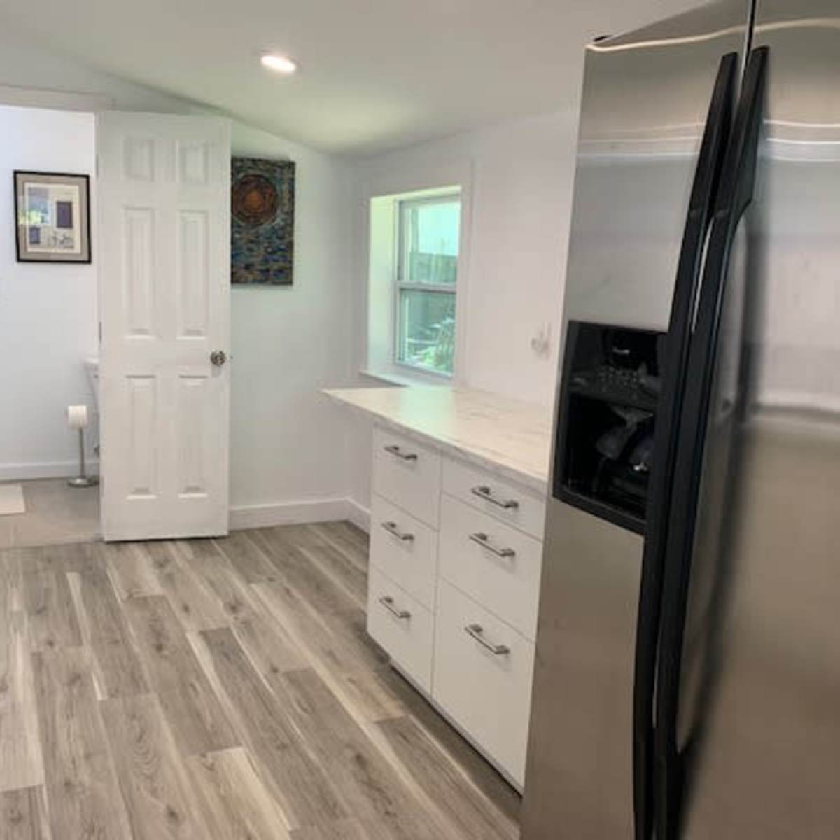 A modern kitchen with light-colored cabinets, a stainless steel refrigerator, and a doorway leading to a bathroom.