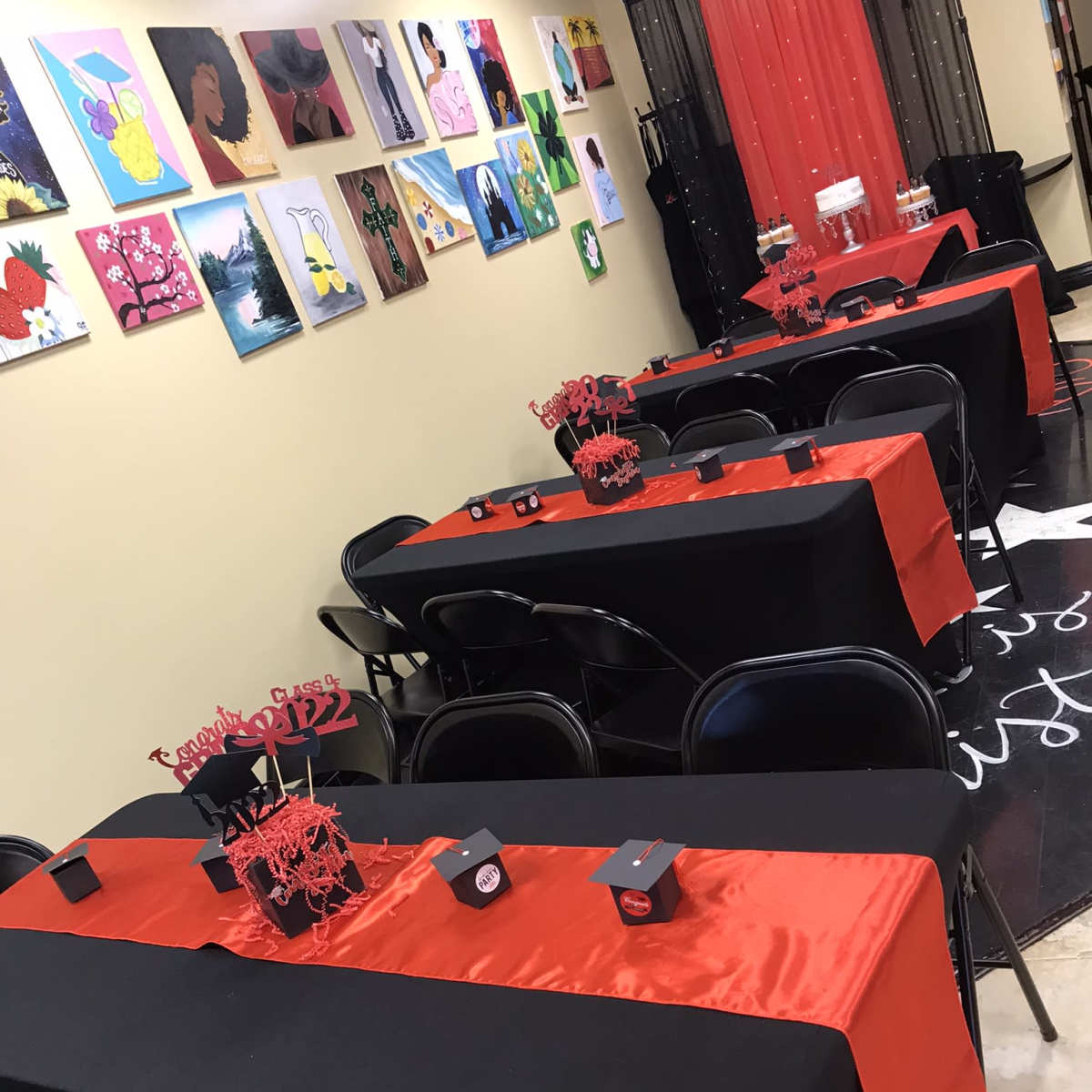 A decorated room with several black tables covered in red table runners, arranged with red centerpieces and surrounded by black chairs, alongside a wall adorned with colorful artworks.