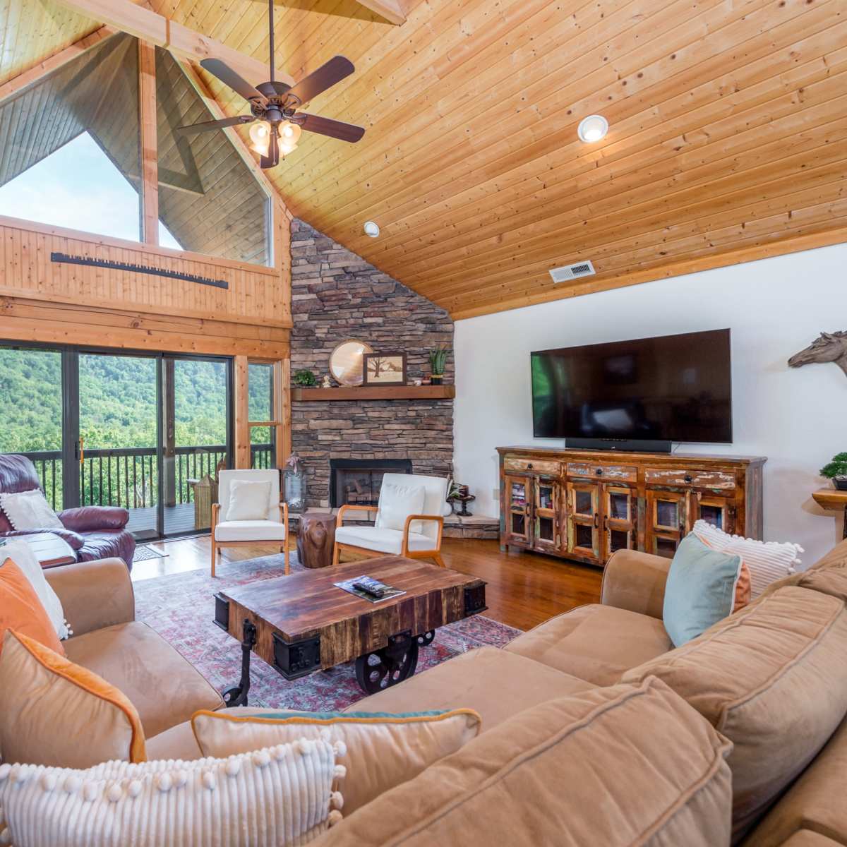 A spacious living room features a large sectional sofa, wooden ceiling beams, a stone fireplace, and a view of lush greenery through large windows.